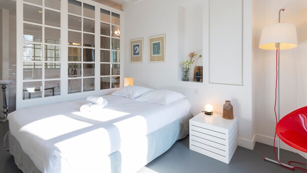 A bedroom with a large bed, a bedside table, a red chair, and a window with a view of the outside.