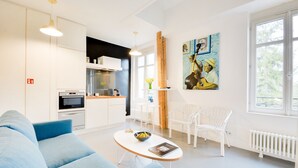 A modern living room with a sofa, a coffee table, and a kitchen area with a stove and cabinets. There are two white chairs and a painting on the wall. - L'Hôtel Particulier - L'appartement de Pierre (Nancy)
