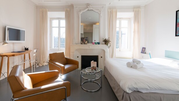 A hotel room with a bed, two armchairs, a small table, a TV, a fireplace, and large windows.