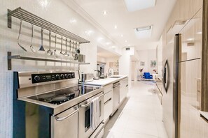 A modern kitchen with stainless steel appliances, a hanging utensil rack, and a view into the adjacent living area. - Uhost AI Miami 5BR Heated Pool Hot Tub (Aventura)