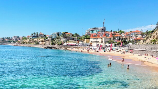 Una playa costera con agua turquesa transparente, una orilla arenosa y edificios al fondo.