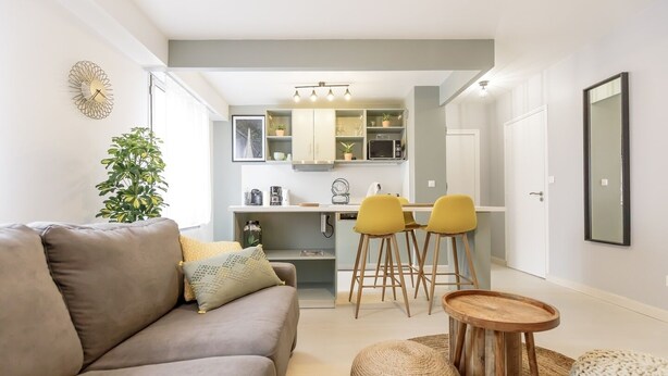 A modern living room with a grey sofa, yellow chairs, a round wooden table, and a kitchen area with a white cabinet and a microwave. - Le Coquet de Simon - Proche de la place Stanislas (Nancy)
