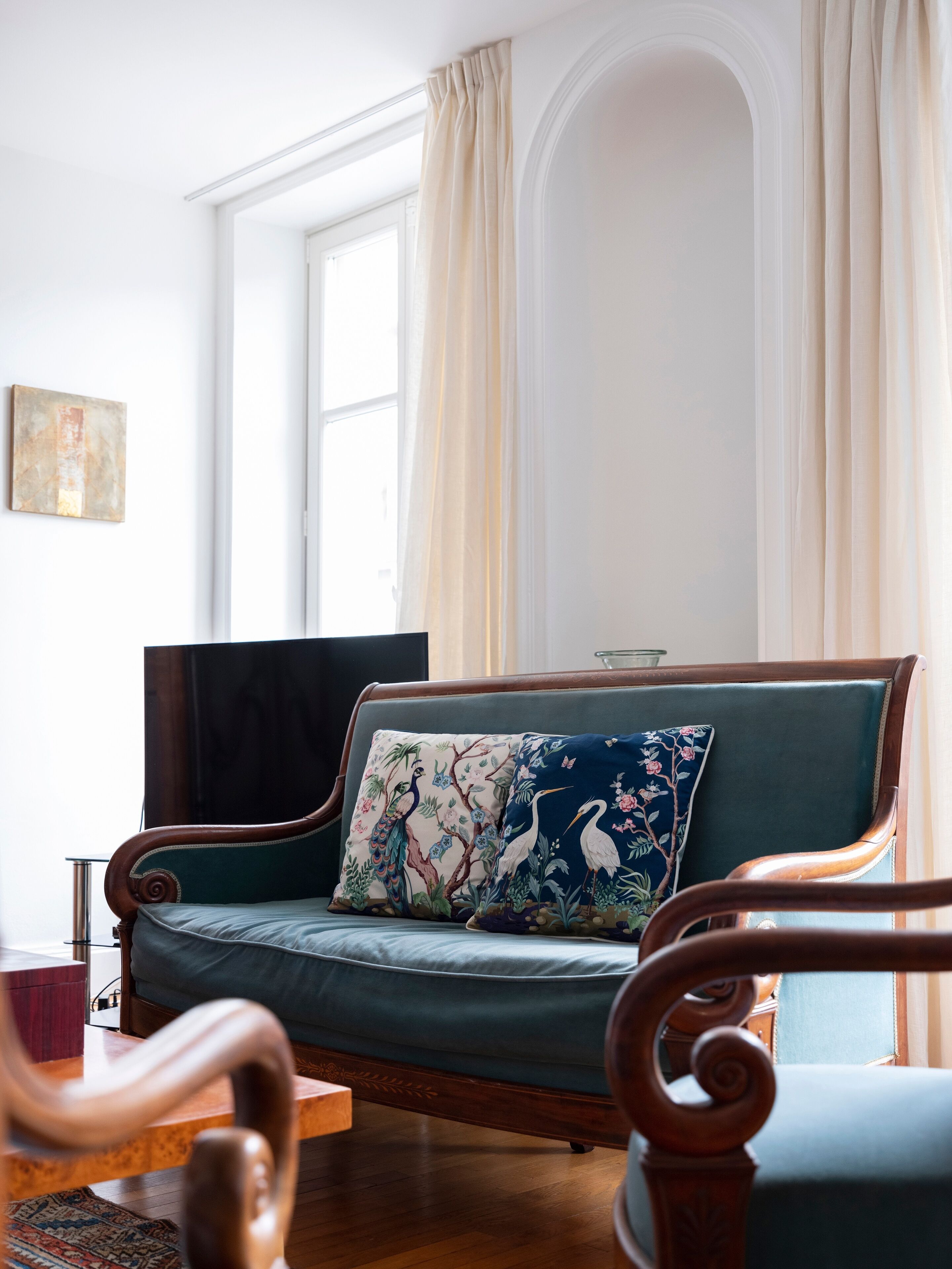 A room with a green sofa, wooden armchairs, a wooden coffee table, and a large window with curtains.
