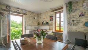 Cozinha rústica com paredes de pedra, uma mesa de jantar de madeira e um vaso de flores, com vista para o verde lá fora.