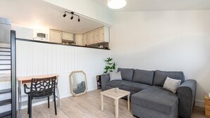 Ein modernes Wohnzimmer mit einem grauen Sofa, einem Holztisch und einem schwarzen Sessel. Im Hintergrund ist ein kleiner Tisch mit einer Mikrowelle und Schränken zu sehen.