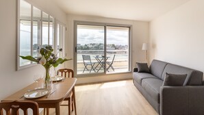 A dining area with a table set for two, a sofa, and a view of the city through large windows. - L'Escale Panoramique - Appt vue mer à Dinard (Dinard)