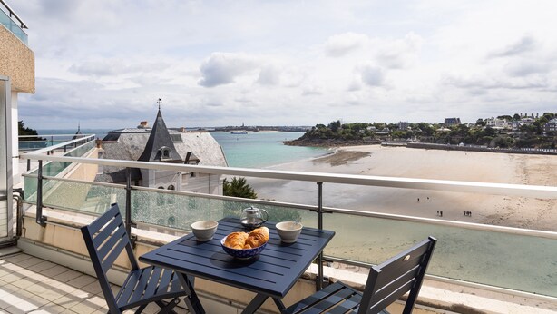 A balcony with a table and chairs, overlooking a beach and a coastal town. - L'Escale Panoramique - Appt vue mer à Dinard (Dinard)