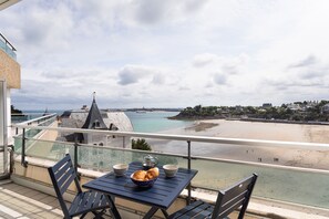 Un balcón con mesa y sillas, con vista a una playa y un pueblo costero.