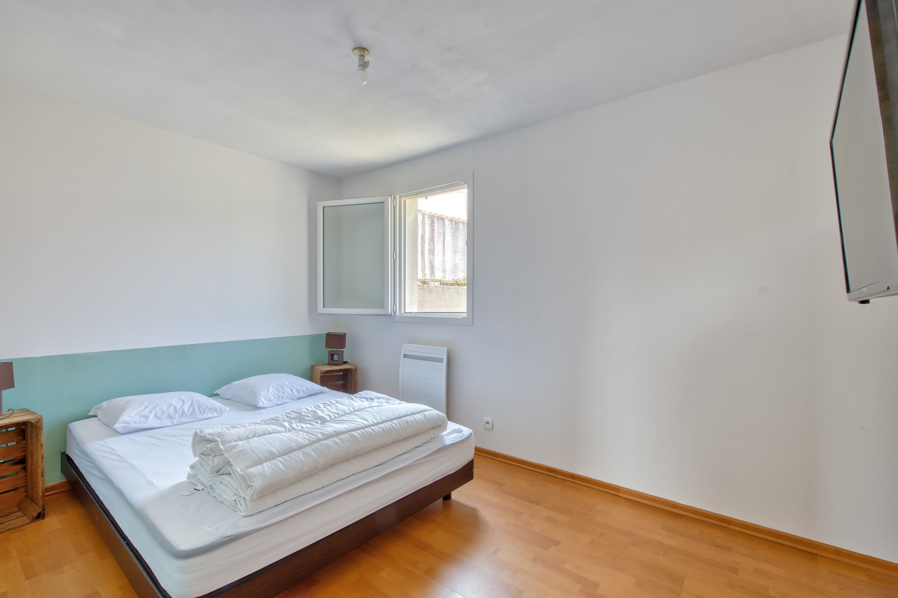 Un dormitorio con cama, suelo de madera, una ventana y una mesita de noche.