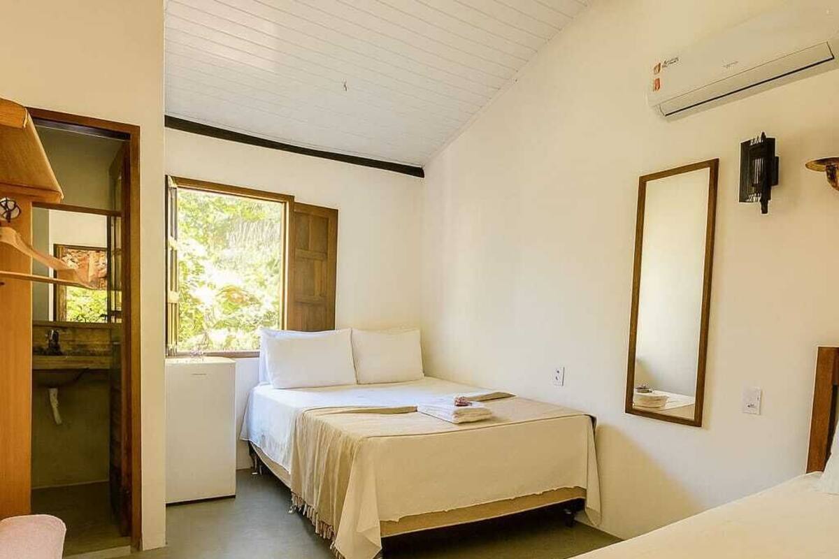 A bedroom with two beds, a wooden wardrobe, a small table, and a window with a view of greenery.