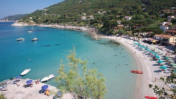 A coastal beach with clear turquoise waters, boats, and beach umbrellas.