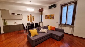 A modern living room with a sectional sofa, a dining table with chairs, a kitchen area with cabinets, and a large window with blinds. - Cozy 1-bedroom apartment in fantastic Milano with AC, WiFi (Milano)