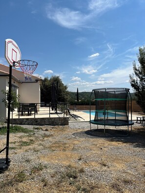 A residential outdoor area with a basketball hoop, a trampoline, a pool, and a patio with chairs and tables.