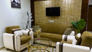 A modern living room with a brown sofa, two armchairs, a glass coffee table, and a flat-screen TV mounted on a textured wall. - Flower Pyramids Inn (Giza)
