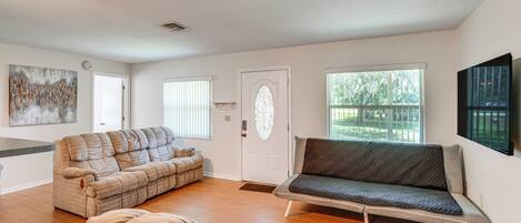 A spacious living room with a large sectional sofa, a smaller sofa, and a bean bag chair. There is a large window providing natural light and a view of the outdoors.
