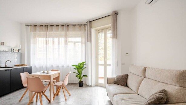 Ein modernes Wohnzimmer mit einem beigen Sofa, einem Esstisch mit Stühlen, einer Zimmerpflanze und einer Küchenzeile mit Spüle und Schränken.