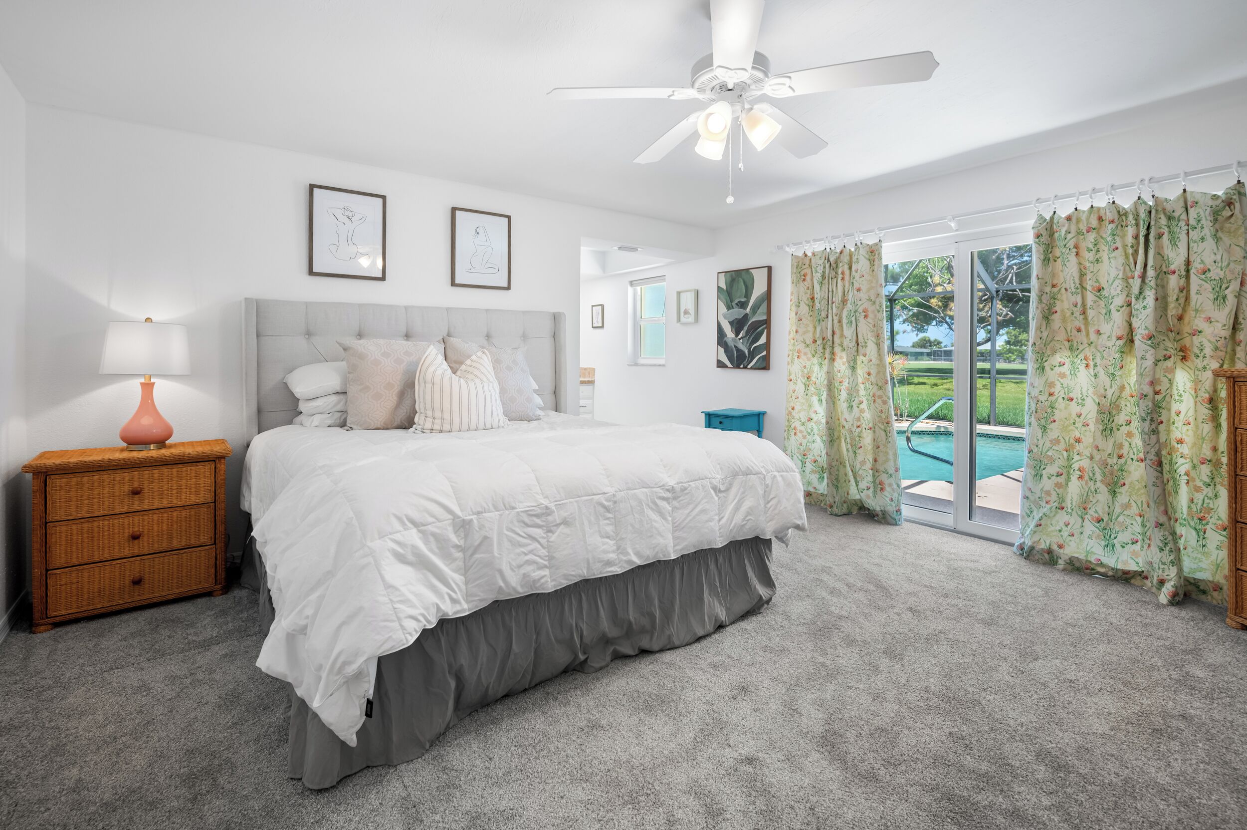 Quarto com cama, mesa de cabeceira, ventilador de teto e vista para uma piscina através de amplas janelas.