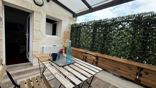 An outdoor patio with a wooden table and benches, a glass bottle, and a vase with flowers.