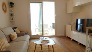 Un salon moderne avec un canapé, une table basse et un téléviseur. Une porte-fenêtre coulissante donne accès à un balcon aménagé avec du mobilier d’extérieur.