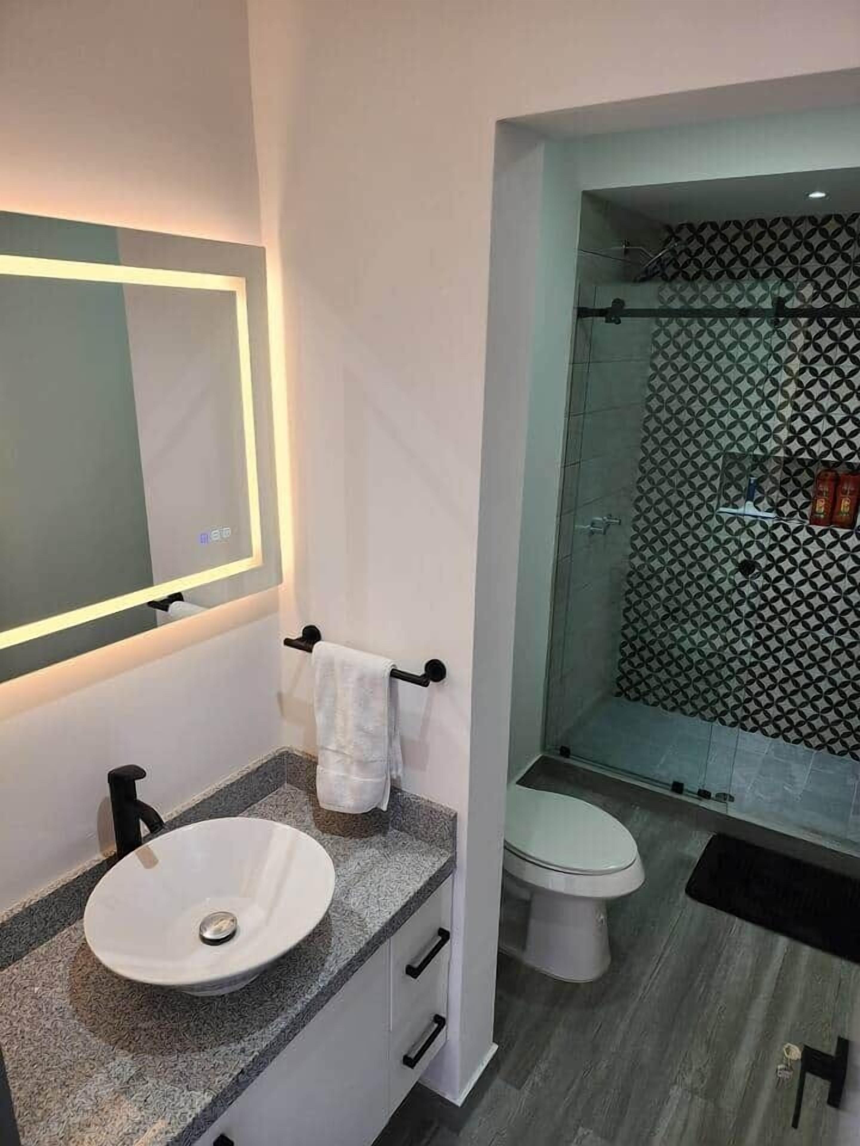 Un baño moderno con un lavabo blanco, un espejo con luz, un perchero, un inodoro y una ducha con detalles decorativos en la pared.
