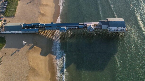 Vista aérea de uma área costeira com uma extensa faixa de areia, um píer que se estende sobre a água e edifícios ao longo da orla.