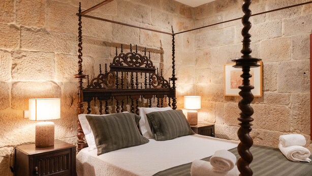 A four-poster bed with a stone wall backdrop, two bedside tables with lamps, and a neatly made bed with a white comforter and green striped pillows. - Feel Discovery Casa da Granja I (Resende)