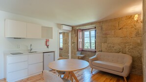 Un espace de vie compact comprenant un coin cuisine, une table de salle à manger ronde en bois, un canapé beige et un mur en pierre.