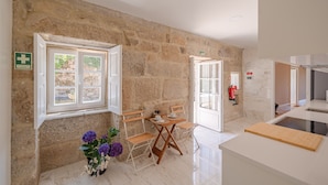 Une pièce aux murs de pierre, une fenêtre ornée d’une croix, une table avec des chaises, un vase rempli de fleurs et un extincteur.