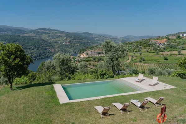 A rectangular swimming pool with lounge chairs and a lifebuoy, surrounded by a grassy area and overlooking a lake and hilly landscape.