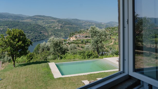Une vue depuis une fenêtre donnant sur une piscine, une végétation luxuriante et des collines lointaines.