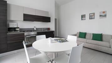 Una cocina moderna con armarios de madera oscura, estufa de acero inoxidable y microondas. Hay una mesa de comedor con sillas blancas y una revista. Un sofá gris con cojines verdes está apoyado contra la pared.