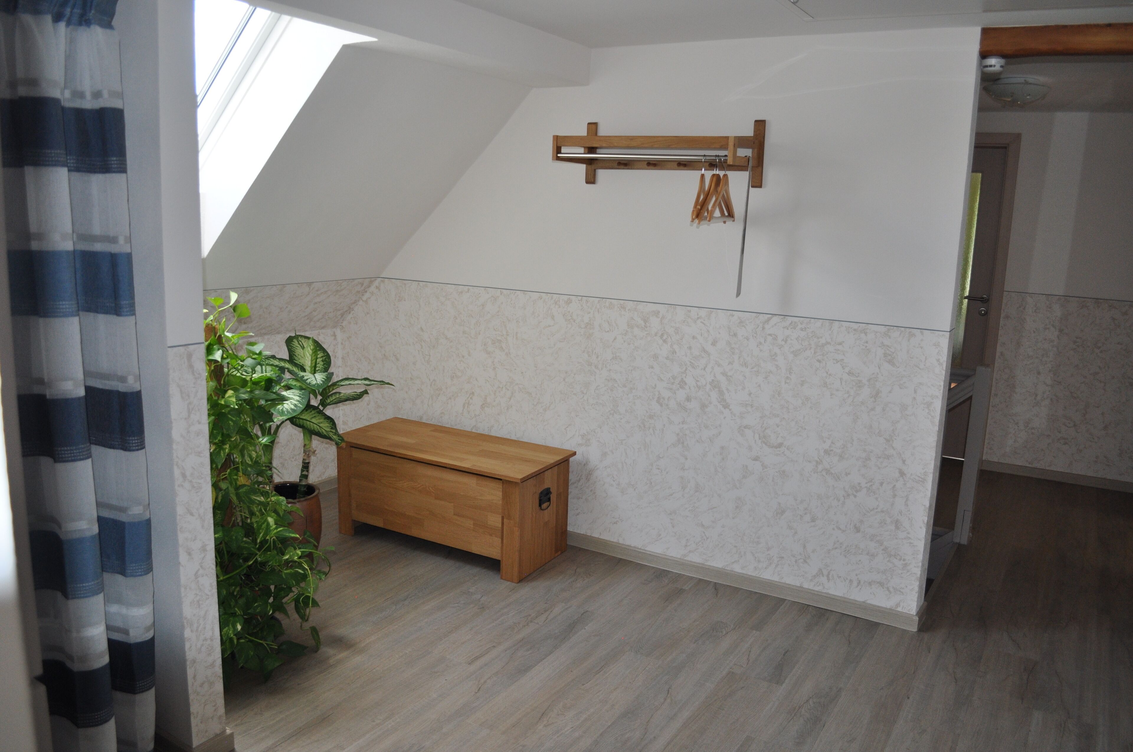 A room with a sloped ceiling, a wooden bench, a hanging coat rack, and a potted plant.