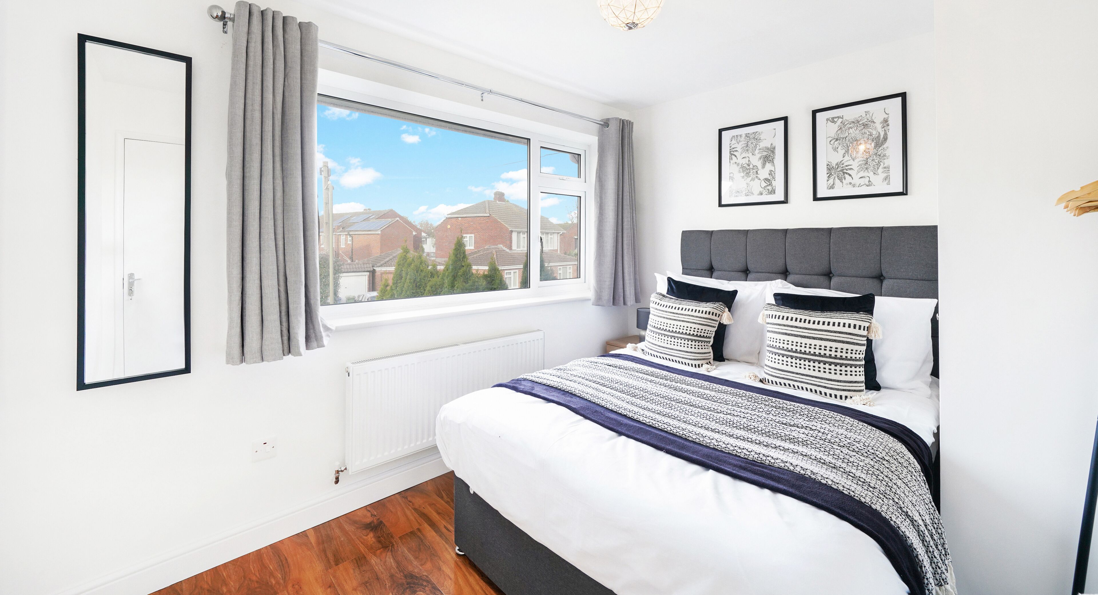 A bedroom with a large window, a bed with patterned pillows, and two framed artworks on the wall.