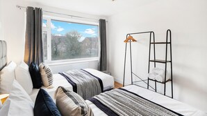 A bedroom with a bed, a window with curtains, a metal shelf with towels, and a view of houses outside. - Brabon Road House By REDWOOD STAYS (Farnborough)