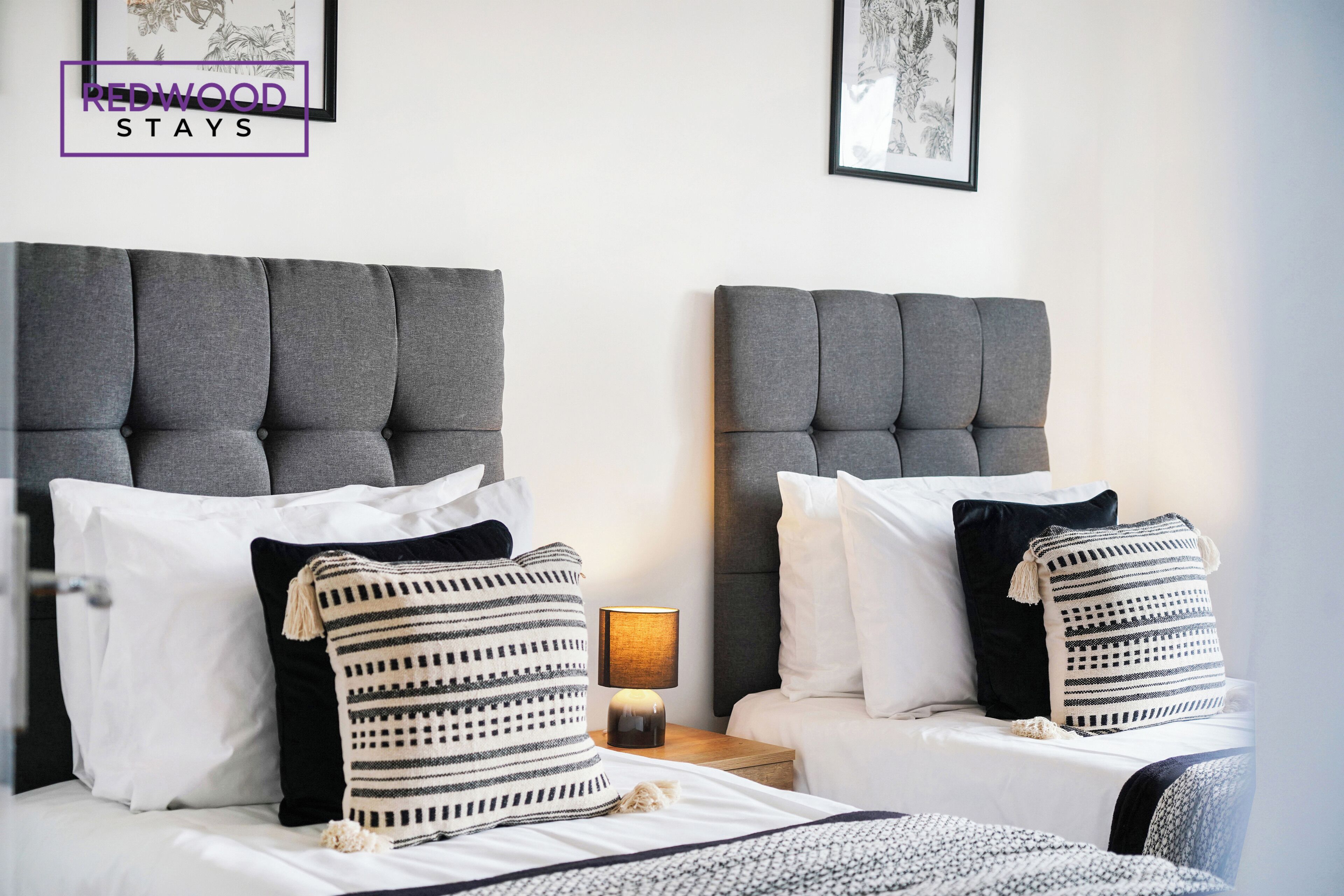 A hotel room with two beds, each with a grey upholstered headboard, white bedding, and striped pillows. A small table lamp is visible between the beds.