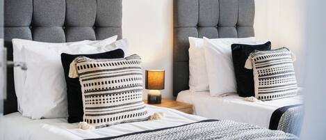 A hotel room with two beds, each with a grey upholstered headboard, white bedding, and striped pillows. A small table lamp is visible between the beds.
