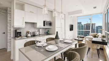 A modern kitchen with white cabinetry, a dining area with a glass table, and a city view through large windows.