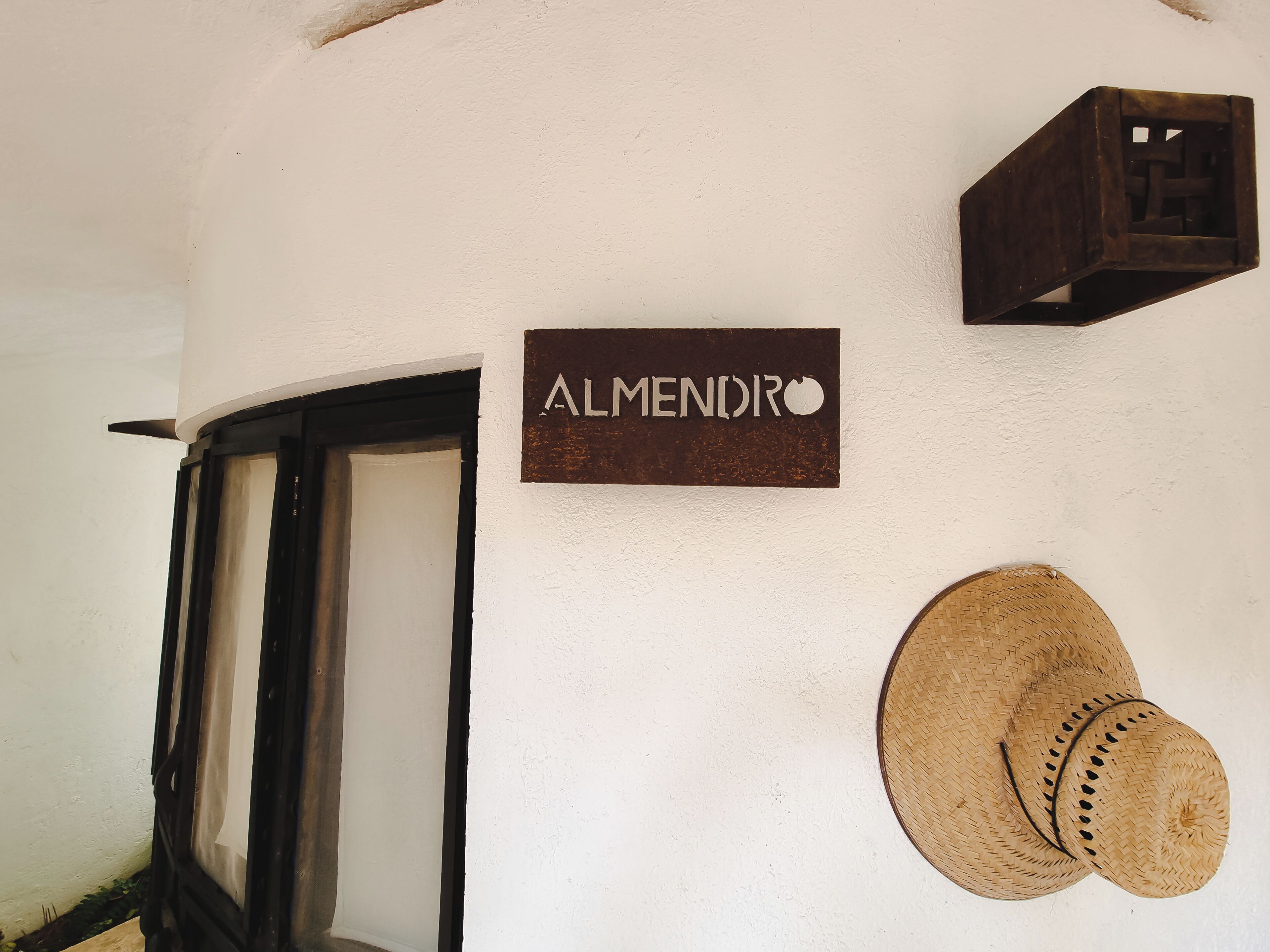 Une enseigne indiquant « ALMENDRO » sur un mur blanc, une caisse en bois et deux chapeaux tressés.