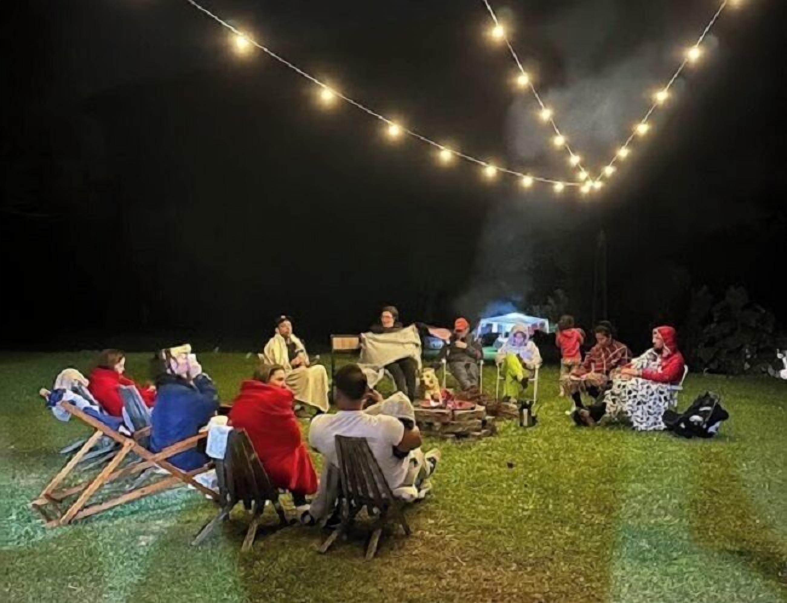 Een groep mensen zitten 's avonds buiten op stoelen, met lichtslingers erboven.