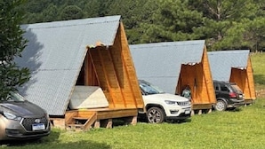 A row of A-frame cabins with metal roofs, parked on a grassy field with a forested hill in the background. - Aldeia Colibri (Jaquirana)