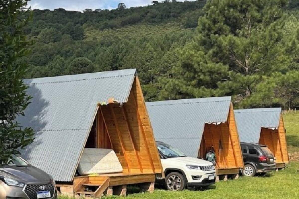 Une rangée de chalets en forme de «A» avec des toits métalliques, garés sur un champ herbeux, avec une colline boisée en arrière-plan.