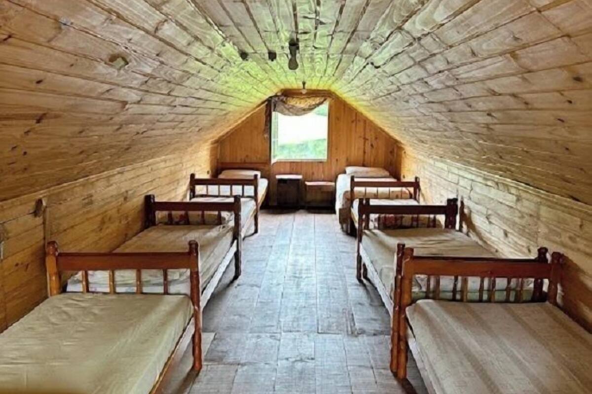 Une longue pièce étroite aux murs en bois et au plafond en pente, contenant quatre lits en bois alignés le long de l’axe central.