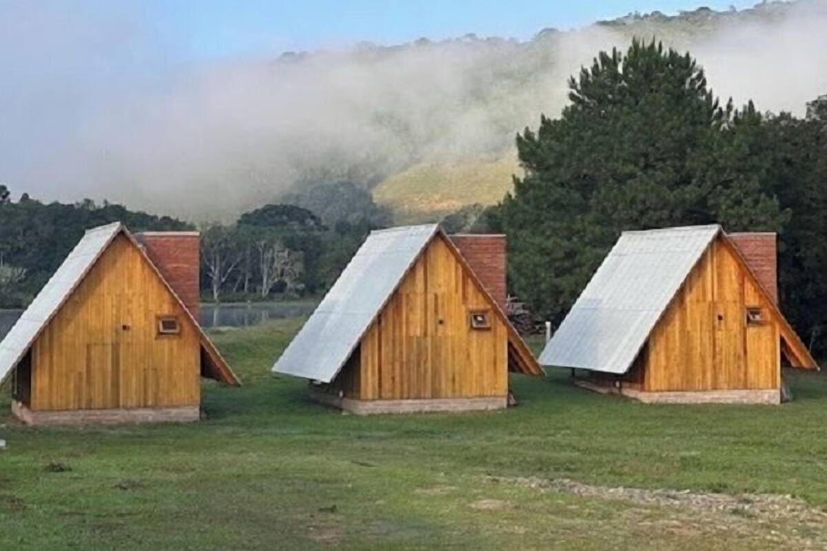 Trois cabanes en bois aux toits en pente, nichées dans un champ verdoyant, avec en arrière-plan des arbres et des collines enveloppées de brume.