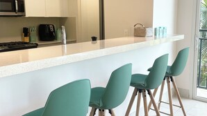 Une cuisine moderne avec des armoires blanches, un four à micro-ondes en acier inoxydable et une machine à café noire. On y trouve trois tabourets de bar verts avec des pieds en bois.