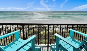 Ein Balkon mit türkisfarbenen Stühlen mit Blick auf einen Strand, der klares Wasser und eine felsige Küste zeigt.