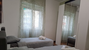 Ein Zimmer mit zwei Betten, jeweils mit einem gemusterten Überwurf und einem aufgerollten Handtuch. Es gibt durchscheinende Vorhänge mit floralem Muster und einen Spiegel an der Wand.