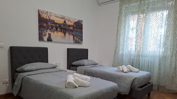 Ein Hotelzimmer mit zwei Betten, grauer Bettwäsche, einem Stadtbildgemälde und einem Fenster mit gemusterten Vorhängen.