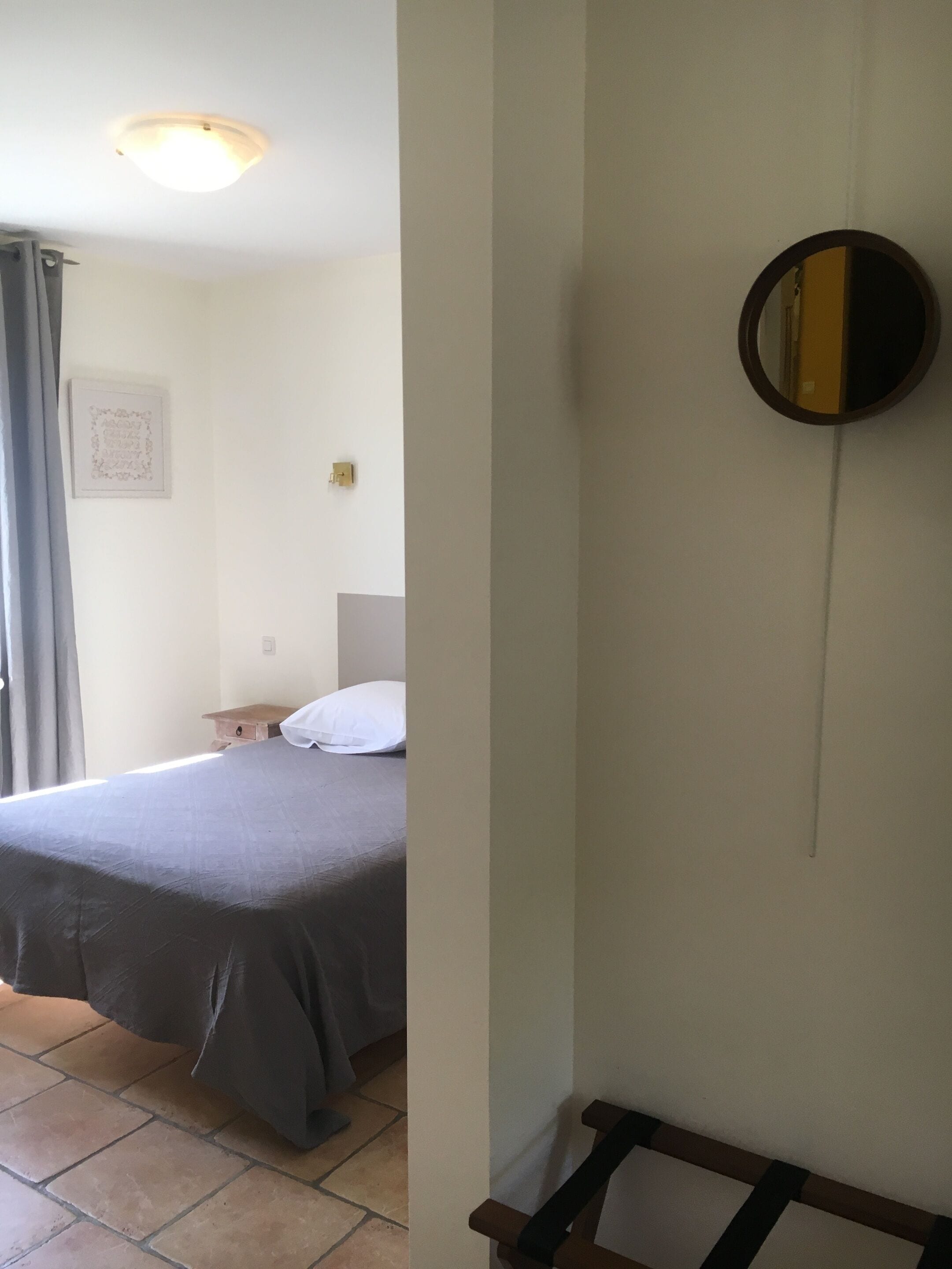 Un dormitorio con cama, mesita de noche, espejo en la pared y ventana con cortinas.
