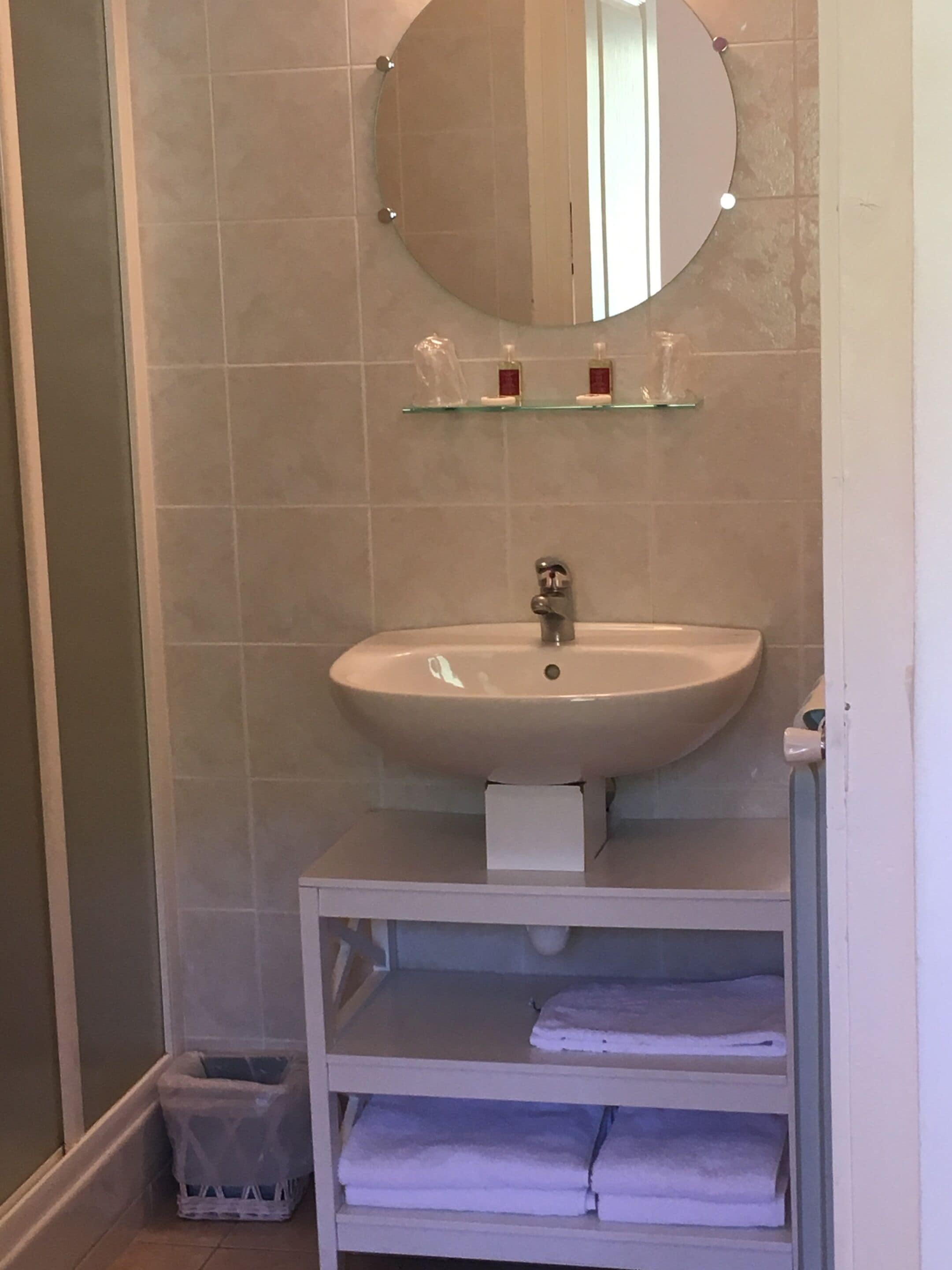 Une salle de bain avec un lavabo blanc, un miroir au-dessus et une étagère en verre où sont rangés les articles de toilette. Un porte-serviettes blanc se trouve sous le lavabo.