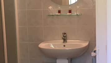 Une salle de bain avec un lavabo blanc, un miroir au-dessus et une étagère en verre où sont rangés les articles de toilette. Un porte-serviettes blanc se trouve sous le lavabo.
